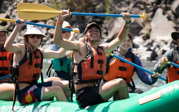 a group of outward bound students sitting in a raft lift their paddles into the air and smile